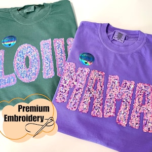 Può includere: Una t-shirt verde salvia con la parola LOLLIE in paillettes rosa e bianche e una t-shirt viola con la parola MAMA in paillettes rosa e bianche. Entrambe le magliette presentano un piccolo adesivo olografico circolare e la scritta Premium Embroidery.