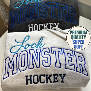 Puede incluir: Dos sudaderas con capucha, una gris oscuro y otra gris claro, están apiladas. Cada una presenta el texto "Lock Monster Hockey" en bordado azul. La sudadera gris claro tiene una pegatina circular que dice "Premium Quality Super Soft".