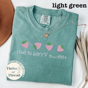 May include: Light green t-shirt with the text "Life is berry sweet" and embroidered pink strawberries. A silver necklace with a pendant is on the shirt. The shirt is labeled "Comfort Colors" and "Custom Embroidery".