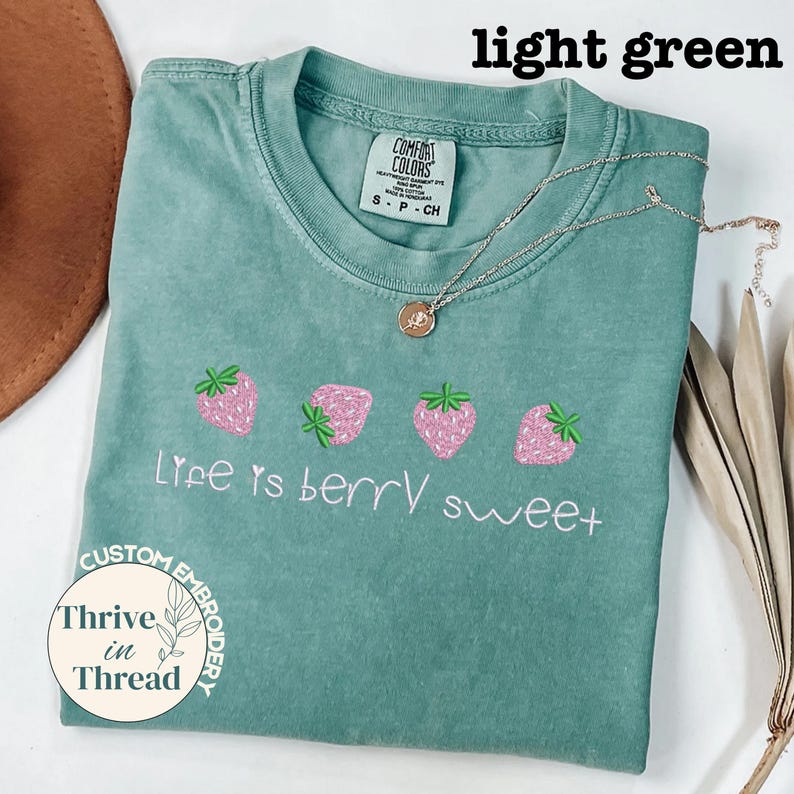 May include: Light green t-shirt with the text "Life is berry sweet" and embroidered pink strawberries. A silver necklace with a pendant is on the shirt. The shirt is labeled "Comfort Colors" and "Custom Embroidery".
