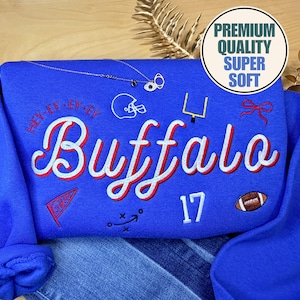 May include: A royal blue sweatshirt featuring the word "Buffalo" in white and red script. The number "17" and a football are embroidered below. Additional embroidered details include a football helmet, goal post, and a red pennant.