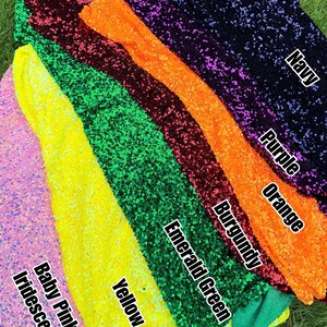 May include: A collection of sequin fabric swatches in various colors, labeled with text: Baby Pink Iridescent, Yellow, Emerald Green, Burgundy, Orange, Purple, and Navy. Each fabric piece is covered in small, reflective, circular sequins.