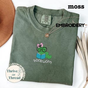 Könnte beinhalten: Moosgrünes T-Shirt mit einem gestickten Bücherwurm-Design. Das Design zeigt einen grünen Wurm mit Brille und rosa Schleife, der ein blaues Buch liest. Der Text "bookworm" steht darunter.