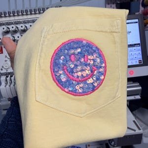 May include: A pale yellow t-shirt featuring a chest pocket adorned with a circular patch. The patch displays a smiley face design filled with iridescent light blue sequins and outlined with a vibrant pink embroidered border.