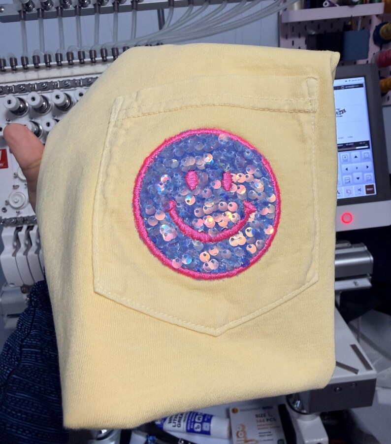 May include: A pale yellow t-shirt featuring a chest pocket adorned with a circular patch. The patch displays a smiley face design filled with iridescent light blue sequins and outlined with a vibrant pink embroidered border.
