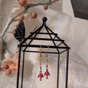 Könnte beinhalten: Ein Paar goldene Ohrhänger mit rosa Pilz-Anhängern und lila und weißen Perlen.