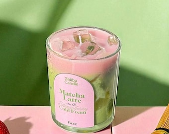 May include: A glass candle shaped like a drink with green matcha liquid, pink strawberry cold foam, and clear ice cubes on top. The label reads Shiba Candie, Matcha Latte with Strawberry Cold Foam, 6oz. Two strawberries sit nearby.