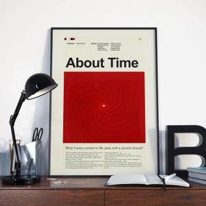 May include: A framed movie poster for "About Time" with a black frame. The poster features a red spiral design with a white heart in the center. The title "About Time" is in bold black letters. A black desk lamp and open book are in the foreground.