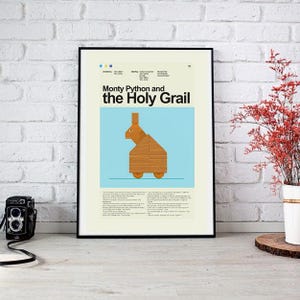 Könnte beinhalten: Ein gerahmtes Poster mit dem Titel "Monty Python and the Holy Grail". Das Poster zeigt eine Holzkaninchen-Illustration auf hellblauem Hintergrund. Das Poster ist in einem schwarzen Rahmen und wird an einer weißen Backsteinmauer gezeigt.