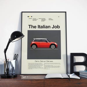 May include: A framed poster for "The Italian Job" film, showing a red Mini Cooper illustration. The poster includes the tagline "Get in. Get out. Get even." and credits. A black desk lamp and open notebook are in the foreground.