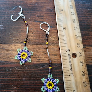 May include: A pair of beaded earrings with a flower design. The earrings are made with purple, green, yellow, and brown beads. The earrings are about 2 inches long.