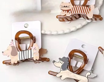 Könnte beinhalten: Vier braune und beige Haarspangen mit Cartoon-Tierdesigns. Eine Spange zeigt eine Katze mit gestreiftem Hemd, eine andere einen Hund mit Partyhut und eine dritte einen Hund mit Hut. Die Spangen befinden sich auf weißen Karten.