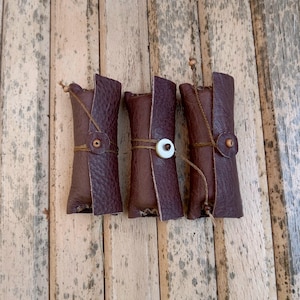 May include: Three cylindrical dark brown leather pouches, each secured with a thin cord wrapped around the center. Two pouches feature a small circular leather tab with a bead, while the middle pouch is fastened with a white circular button.