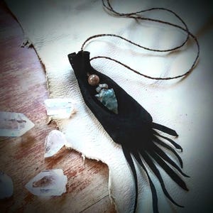 May include: A black suede pendant necklace with a leather cord. The pendant features a small, light blue stone and a brown bead. Clear quartz crystals are scattered around the necklace on a wooden surface.