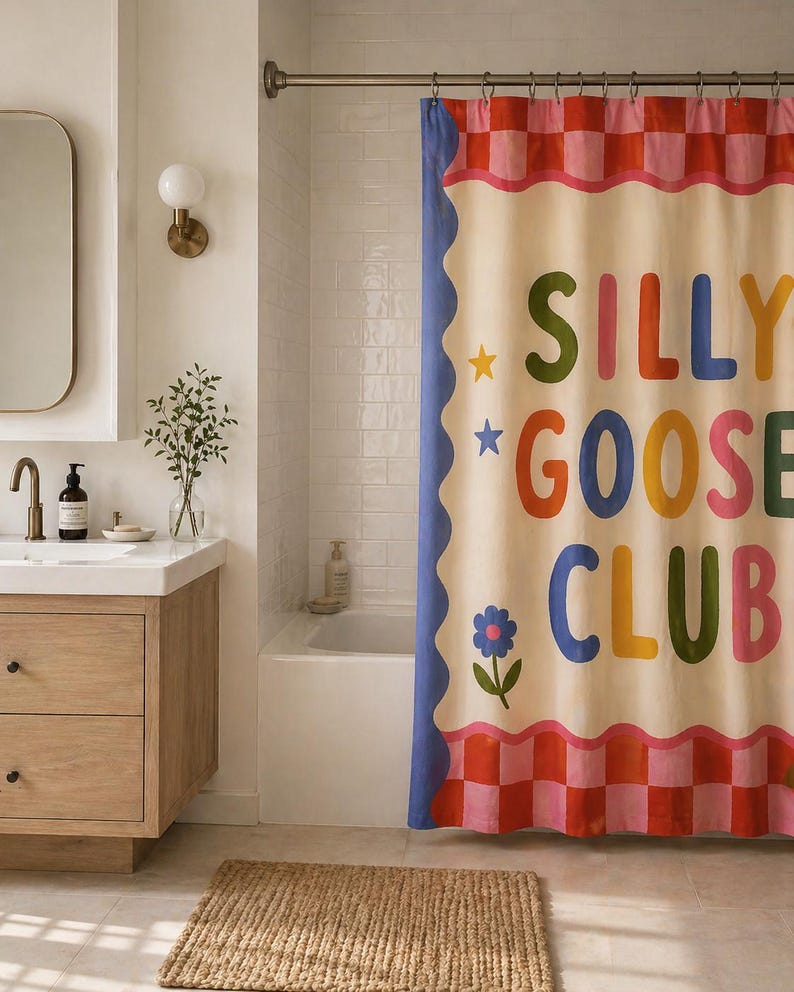 May include: A shower curtain featuring a cream-colored center with the text SILLY GOOSE CLUB in multicolored letters. The design includes a blue scalloped border, small star and flower icons, and a pink and red checkered pattern at the top and bottom.