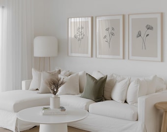 May include: A white sectional sofa with several beige and one olive green throw pillow, a round white coffee table with a small vase of dried grass, and three framed wall art prints featuring black line drawings of flowers on a beige background.