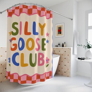 May include: A shower curtain featuring a cream-colored center with the text SILLY GOOSE CLUB in multicolored bubble letters. The design includes small star and flower accents, framed by a wavy pink border and a red and pink checkered pattern.