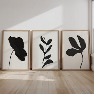Può includere: Tre stampe artistiche incorniciate con disegni floreali neri minimalisti su sfondo crema. Le stampe sono in cornici di legno chiaro e sono esposte su un pavimento in legno. L'opera d'arte comprende forme astratte di fiori e foglie.