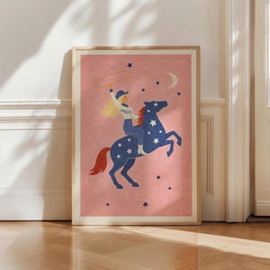 May include: A framed art print featuring a cowgirl riding a blue horse adorned with white stars. The cowgirl, with blonde hair and a blue hat, is throwing a lasso. The background is a soft pink with a crescent moon and scattered stars.