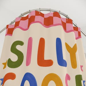 May include: A cream-colored shower curtain hanging from a silver rod, featuring a top border of pink and orange checkered squares. The curtain displays the text SILLY GOOSE in large, multi-colored, rounded letters, accented by small star shapes.