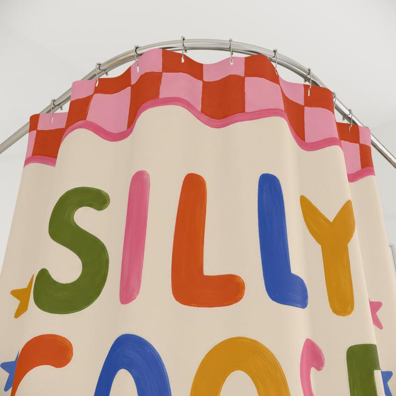 May include: A cream-colored shower curtain hanging from a silver rod, featuring a top border of pink and orange checkered squares. The curtain displays the text SILLY GOOSE in large, multi-colored, rounded letters, accented by small star shapes.