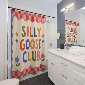 May include: A shower curtain featuring a cream background with a pink and red checkered border at the top and bottom. The text SILLY GOOSE CLUB is printed in large, colorful, rounded letters, surrounded by small star and flower illustrations.