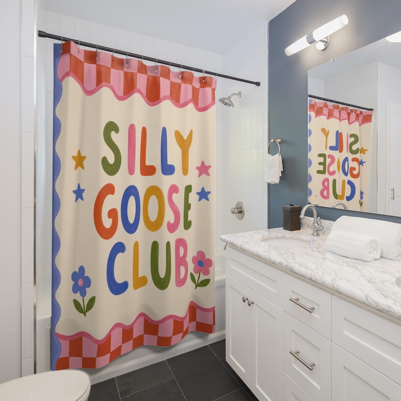 May include: A shower curtain featuring a cream background with a pink and red checkered border at the top and bottom. The text SILLY GOOSE CLUB is printed in large, colorful, rounded letters, surrounded by small star and flower illustrations.