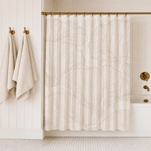 May include: A white shower curtain featuring a large-scale, light beige line art pattern of overlapping fan-shaped leaves. The curtain hangs from a gold-toned rod with matching rings, positioned in a bathroom with white vertical wall paneling.