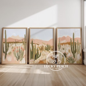 Può includere: Set di tre stampe artistiche incorniciate raffiguranti un paesaggio desertico. L'opera d'arte raffigura cactus, montagne e flora desertica in tonalità di verde, pesca e beige. Le cornici sono in legno chiaro e le stampe sono esposte su un pavimento in legno.