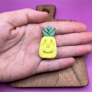 May include: A yellow pineapple with a carved jack-o'-lantern face. The pineapple has green leaves on top.