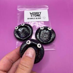May include: Three black worry stones with white googly eyes. The stones are covered in glitter and have a small indentation in the centre. The stones are packaged in a clear plastic bag with a label that reads "Worry Stone Sparkle Bloop".