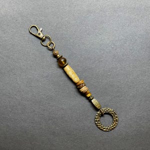 May include: A brown and gold beaded keychain with a round, textured metal charm. The keychain features a variety of beads, including a long, cylindrical bead with a white pattern.