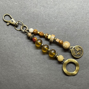 May include: A brown and gold beaded keychain with a world map charm and a circular charm. The beads are a mix of brown, gold, and white.