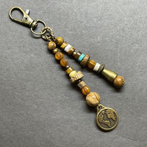 May include: A brown and gold beaded keychain with a world map charm. The beads are made of various materials, including wood, stone, and glass. The keychain is attached to a metal ring.