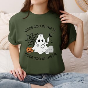 Puede incluir: Camiseta verde oliva con un diseño de Halloween. El diseño presenta un fantasma con gorro de enfermera y estetoscopio, una calabaza, una lápida y el texto "CODE BOO IN THE ER". También se incluye una telaraña y una luna creciente.