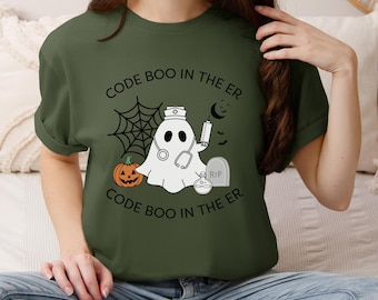 Pode incluir: T-shirt verde-oliva com um design de Halloween. O design apresenta um fantasma com um chapéu de enfermeira e estetoscópio, uma abóbora, uma lápide e o texto "CODE BOO IN THE ER". Uma teia de aranha e uma lua crescente também estão incluídas.