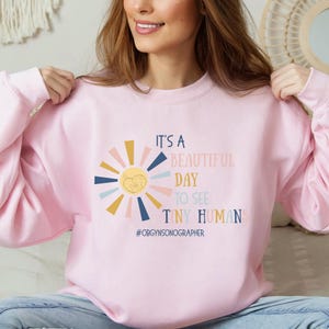May include: Pink sweatshirt with a sun graphic and the text "It's a beautiful day to see tiny humans #OBGYNSONOGRAPHER"
