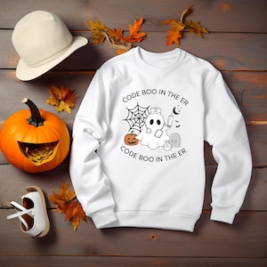 May include: White sweatshirt with a Halloween graphic. The design includes a ghost wearing a stethoscope, a spiderweb, a pumpkin, and the text "CODE BOO IN THE ER". A carved pumpkin and a white hat are also in the image.