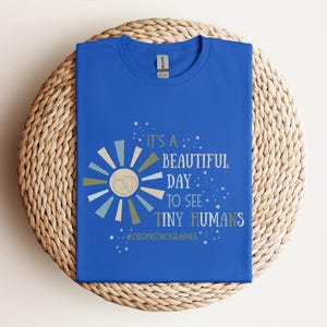 Op de afbeelding: Een koninklijk blauw t-shirt met een afbeelding van een zon met een gestileerde baby erin. De tekst luidt "It's a beautiful day to see tiny humans #obgynSonographer".