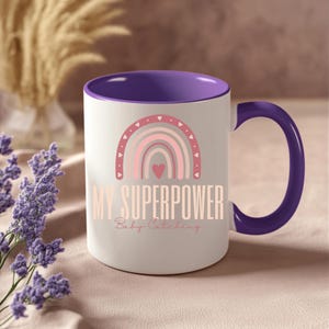 Op de afbeelding: Witte keramische mok met een paars interieur en handvat. De mok heeft een roze en grijs regenboogontwerp met de woorden "MY SUPERPOWER Baby Catching" in een zacht, vrouwelijk lettertype. Een ideaal cadeau voor nieuwe ouders.