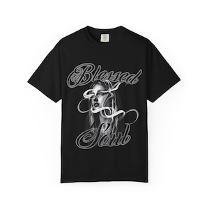 May include: A black short-sleeve t-shirt featuring a graphic print of a person's face partially obscured by swirling white ribbons. The text "Blessed Soul" is printed in a stylised grey cursive font above and below the central portrait image.