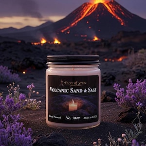 Może przedstawiać: Przezroczysty szklany słoik na świecę z czarną pokrywką i etykietą z napisem Flint & Steel Volcanic Sand & Sage, No. 7049, Hand Poured, Made in the USA. Etykieta przedstawia zapaloną świecę na wulkanicznej skale oraz fioletowe kwiaty.