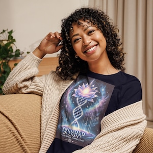 Puede incluir: Una camiseta azul marino con un diseño gráfico que presenta una flor de loto púrpura, una cadena de ADN y un fondo de galaxia. El texto "Encoded in Light" y "Interstellar Expressions" está impreso en la camiseta.