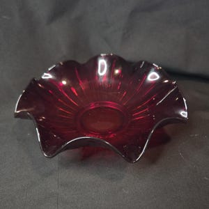 May include: A deep ruby red glass bowl featuring a scalloped, ruffled edge and a series of vertical ribs radiating from the center base toward the rim. The translucent glass has a smooth, polished finish with light reflections on the curved edges.