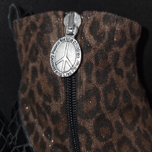 May include: Close-up of a brown and black leopard print boot with a silver zipper. The zipper pull features a peace sign and the text "Donald and Lisa Pliner Peace for the children."