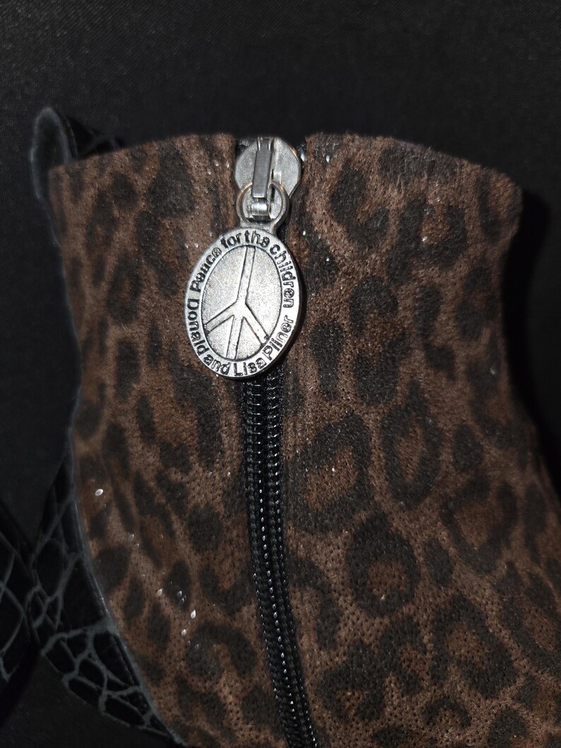 May include: Close-up of a brown and black leopard print boot with a silver zipper. The zipper pull features a peace sign and the text "Donald and Lisa Pliner Peace for the children."