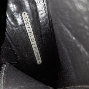 May include: Interior view of a black leather boot. The boot's insole features the brand name "DONALD J PLINER" in white lettering on a rectangular label. The boot's leather appears worn.