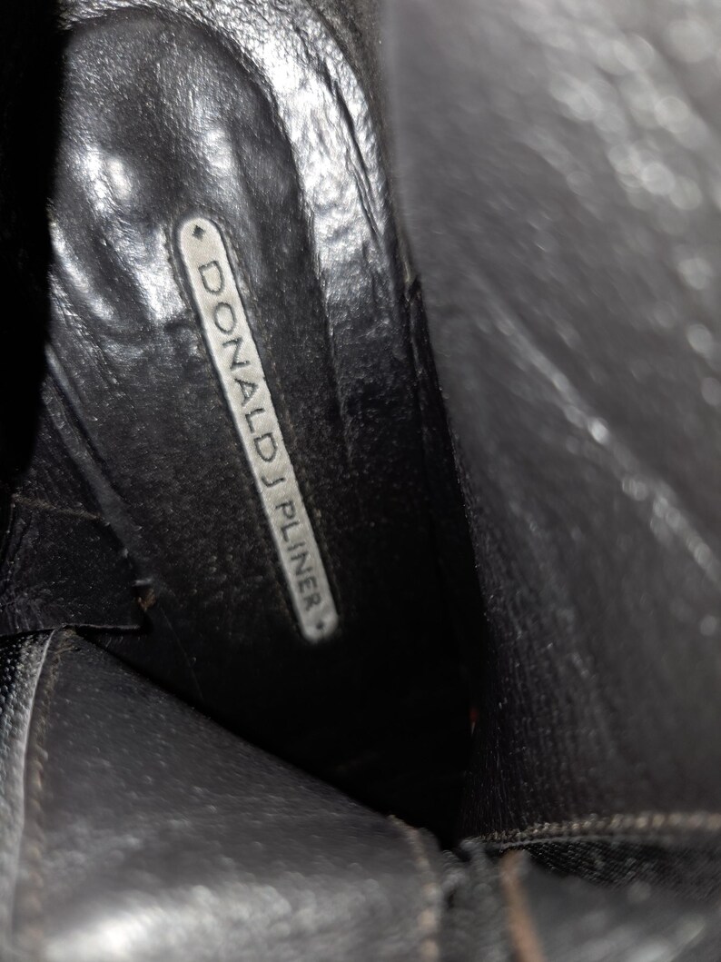 May include: Interior view of a black leather boot. The boot's insole features the brand name "DONALD J PLINER" in white lettering on a rectangular label. The boot's leather appears worn.
