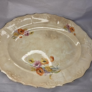 May include: An oval ceramic serving platter with a cream-colored, mottled finish and a scalloped, embossed rim. The platter features two floral arrangements with orange, yellow, and purple flowers and green leaves painted on the surface.