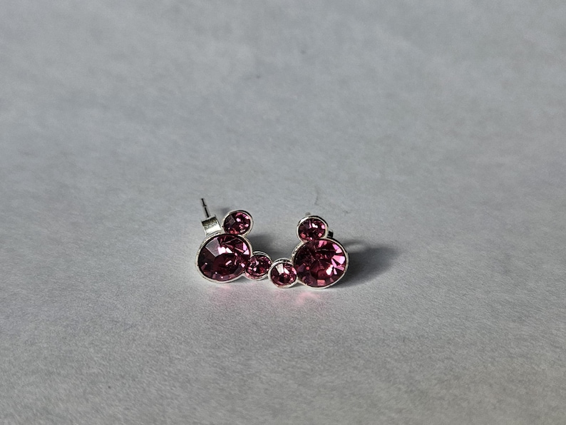 Op de afbeelding: Een paar zilverkleurige oorstekers met een Mickey Mouse-ontwerp. Elke oorbel heeft drie ronde, roze kristallen stenen. De oorbellen zijn geplaatst tegen een effen, lichtgrijze achtergrond.